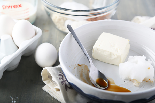 Cream cheese, coconut oil, and vanilla extract in a white bowl with eggs nearby