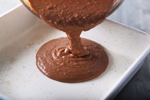 Pouring thick chocolate brownie batter from a glass bowl into a white baking pan