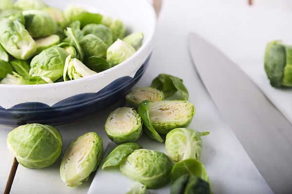 Halved Brussels sprouts on a cutting board with a chef knife and a bowl of whole sprouts