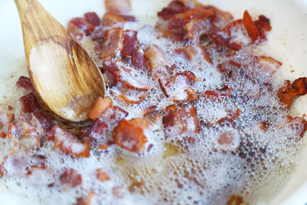 Chopped bacon pieces frying in olive oil with rendered fat bubbling in a pan