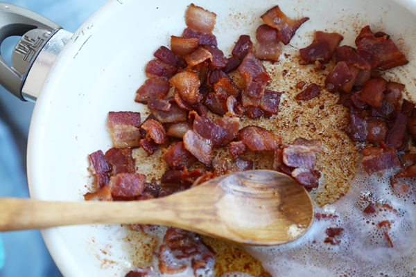 Browned bacon pieces in a white pan with pooling bacon fat and a wooden spoon