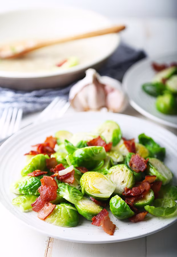 Tender Brussels sprouts topped with crispy bacon on a white plate with garlic behind