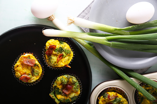 Baked bacon frittata cups with golden edges on a black plate beside green onions