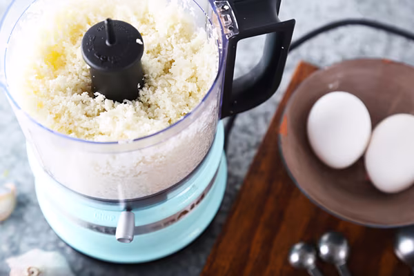 Riced cauliflower in a food processor with eggs on a wooden board nearby