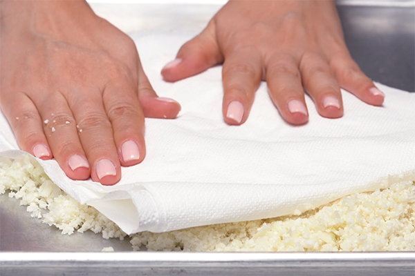 Pressing paper towels over riced cauliflower on a sheet pan to drain moisture
