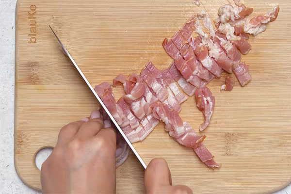 Slicing raw bacon into thin strips on a wooden cutting board