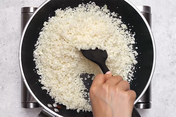 Stirring riced cauliflower with a spatula in bacon fat in a large skillet