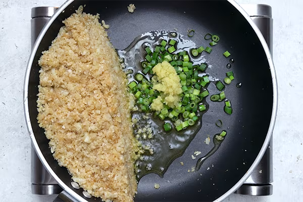 Minced garlic, ginger, and sliced green onions added beside cauliflower rice in a skillet