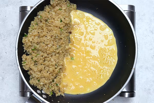 Beaten eggs poured alongside seasoned cauliflower rice in a skillet