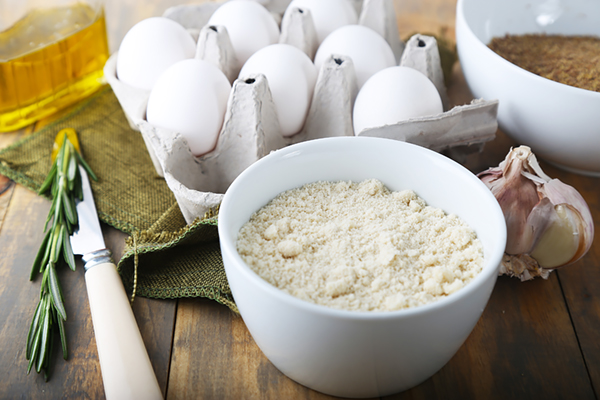 Blanched almond flour in a white bowl with eggs, olive oil, garlic, and rosemary nearby