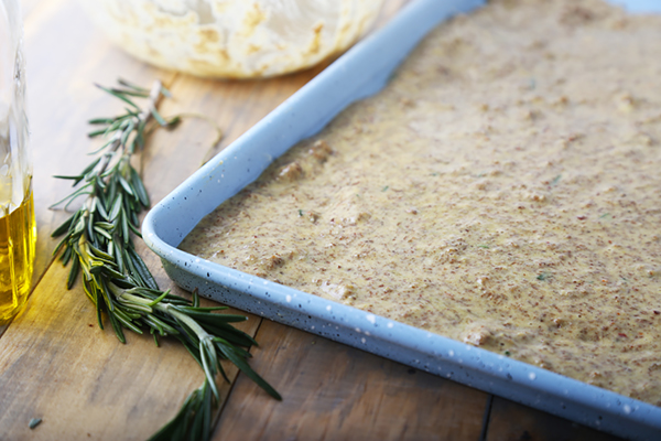 Focaccia batter spread evenly in a blue baking pan with rosemary alongside