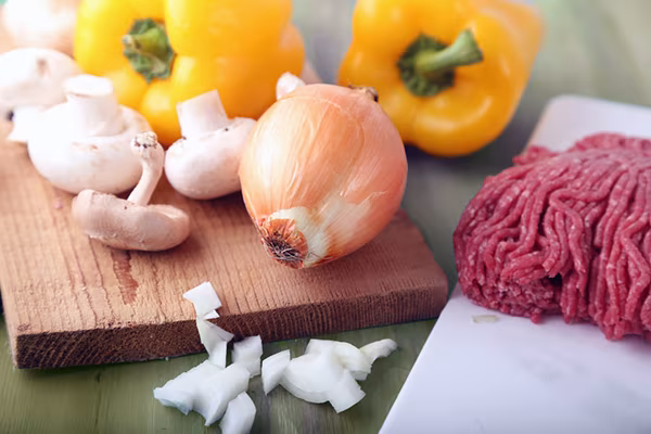 Raw ground beef, whole cremini mushrooms, onion, and yellow bell peppers on a cutting board