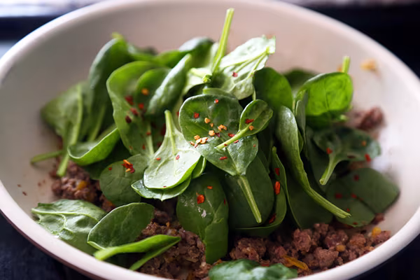 Fresh spinach leaves and red pepper flakes piled over browned ground beef in a white pan