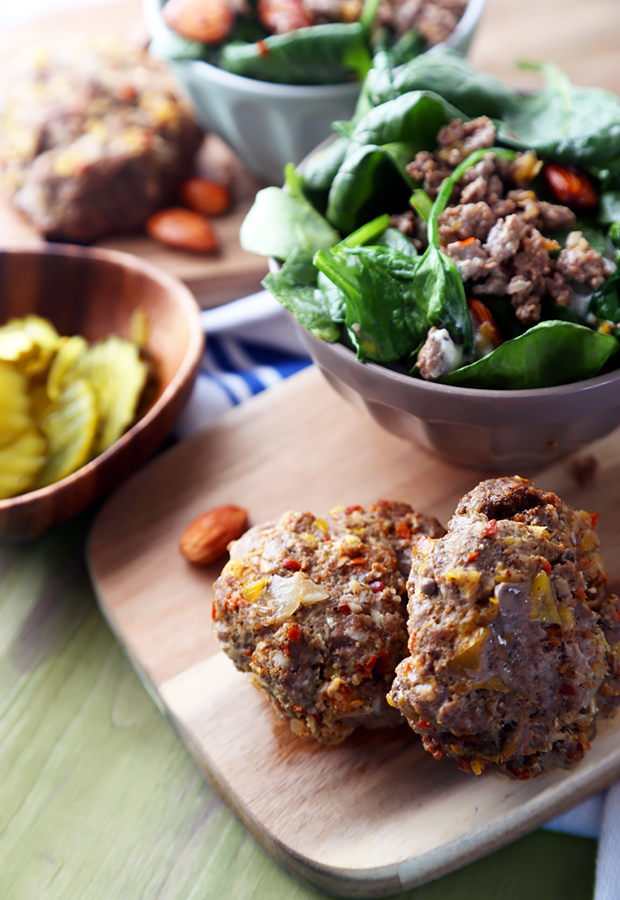 Browned omnivore burgers on a wooden board with creamed spinach, almonds, and pickles alongside
