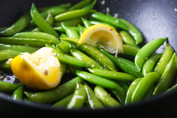Sugar snap peas sauteing with lemon wedges in bacon fat in a dark pan