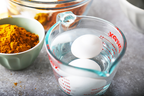 Two eggs submerged in warm water in a glass measuring cup beside curry powder