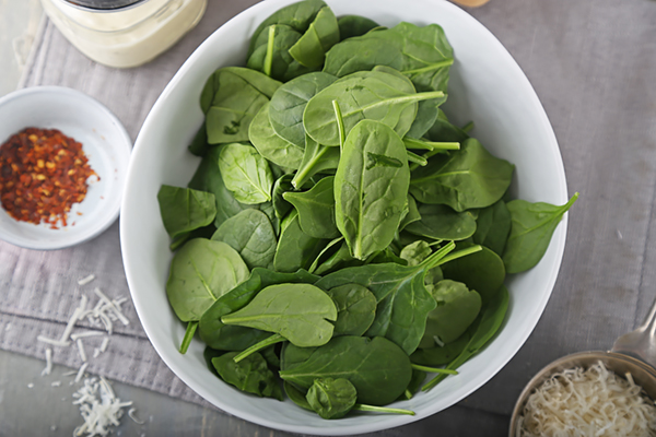 Fresh baby spinach leaves filling a large white bowl with red pepper flakes and parmesan nearby