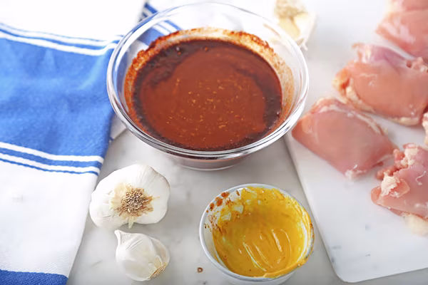 Dark keto BBQ sauce in a glass bowl with yellow mustard, garlic, and raw chicken thighs