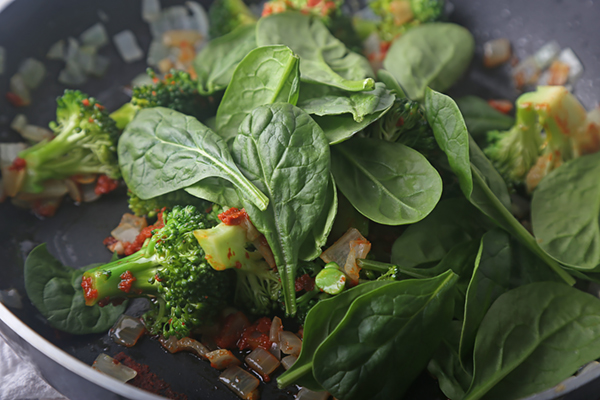 Fresh spinach leaves piled over broccoli and red curry paste in the pan
