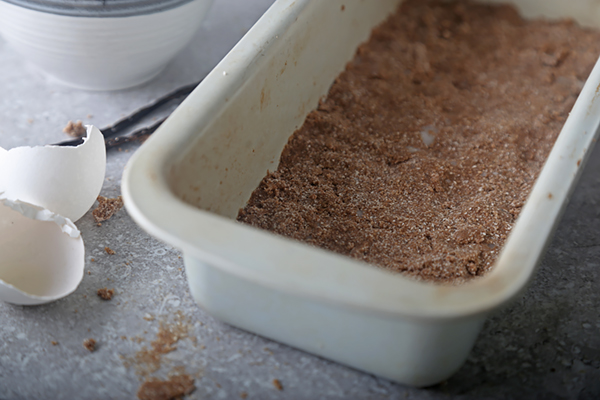 Dark cocoa gingerbread dough pressed into a white loaf pan with eggshell nearby