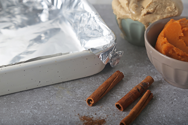 Foil-lined square pan beside pumpkin puree and cinnamon sticks on a counter