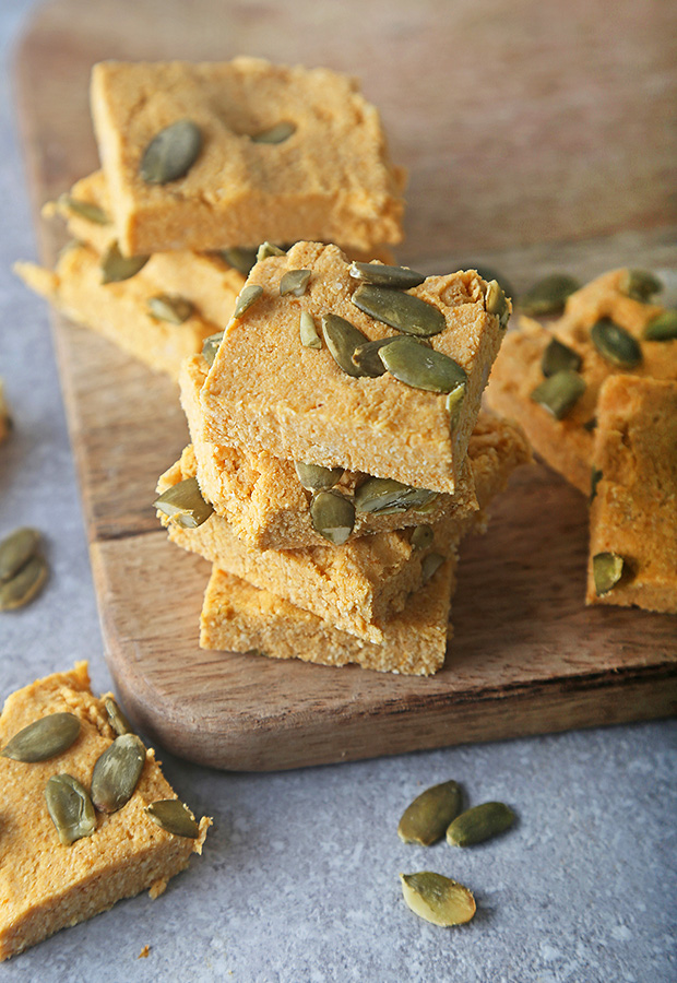 Stacked pumpkin fudge squares topped with pepitas on a wooden cutting board