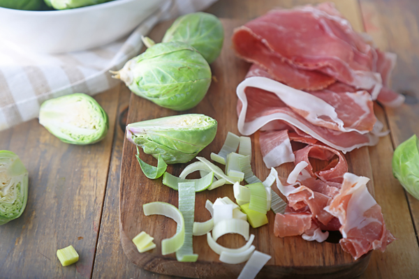 Halved brussels sprouts, sliced leeks, and prosciutto on a wooden cutting board