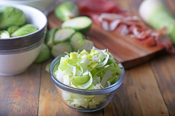 Sliced leeks in a small glass bowl with halved brussels sprouts nearby