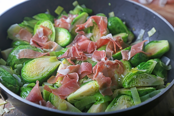 Torn prosciutto scattered over brussels sprouts and leeks in the pan
