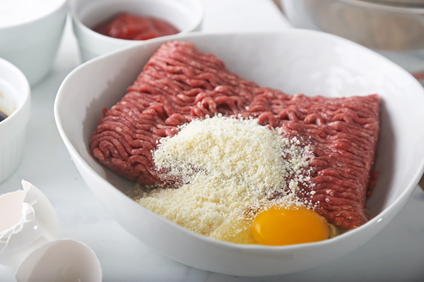 Ground beef, grated parmesan, and a cracked egg in a white mixing bowl