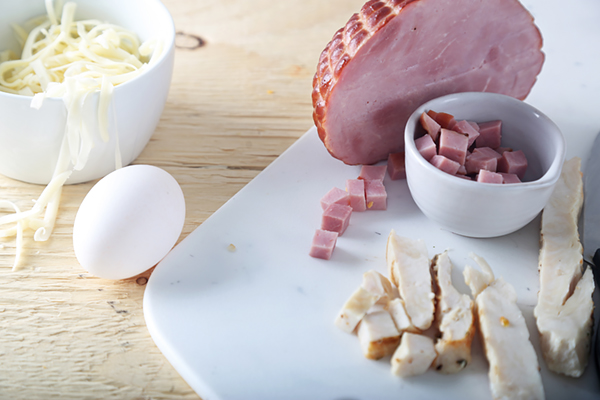 Diced ham, sliced chicken, a whole egg, and shredded cheese on a white cutting board