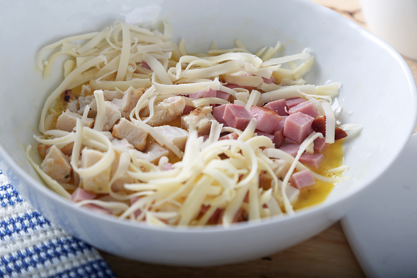 Beaten eggs mixed with diced chicken, cubed ham, and shredded Swiss cheese in a ceramic bowl