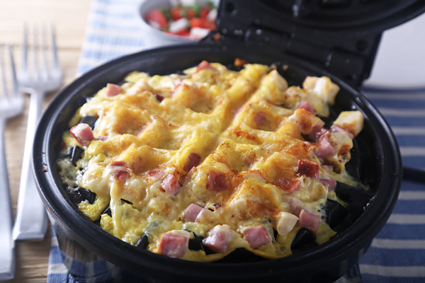 Golden chicken and ham waffle cooking in a black waffle iron with melted cheese