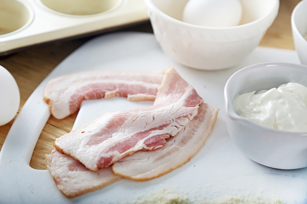 Raw bacon strips on a cutting board next to a small bowl of sour cream and eggs