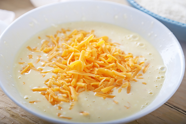 Grated cheddar cheese added to smooth pale muffin batter in a round bowl