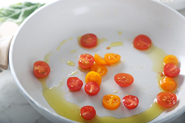 Halved red and yellow cherry tomatoes frying in olive oil in a white pan