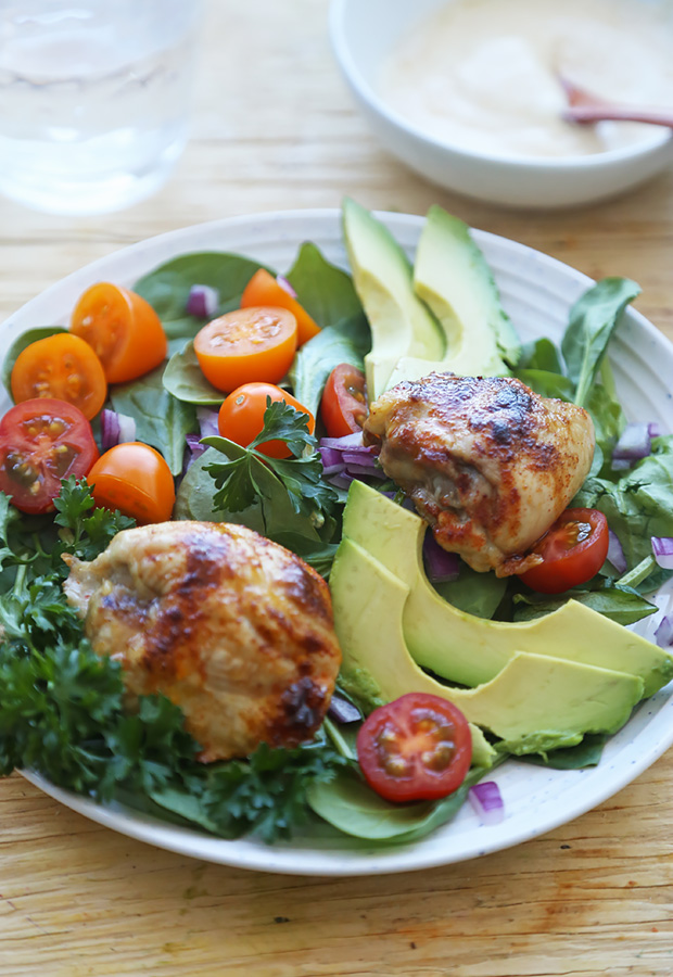 Baked chicken thighs served over greens with avocado slices, cherry tomatoes, and red onion