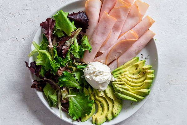 Deli turkey slices, fanned avocado, spring mix, and cream cheese on a white plate