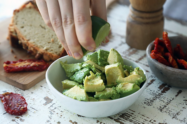 Squeezing lime juice over chunked avocado in a white bowl with sun-dried tomatoes nearby