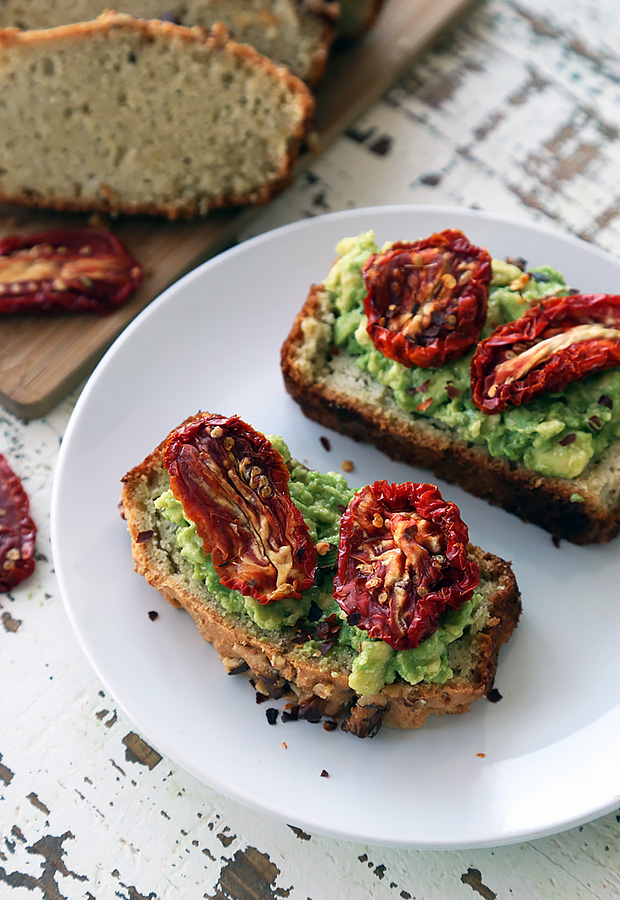 Focaccia toast topped with mashed avocado, sun-dried tomatoes, and red pepper flakes on a white plate