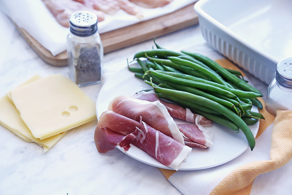 Prosciutto slices, swiss cheese, and fresh green beans arranged on a plate with seasonings