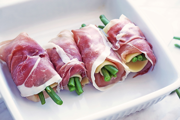 Four prosciutto-wrapped chicken rolls placed seam side down in a white baking dish