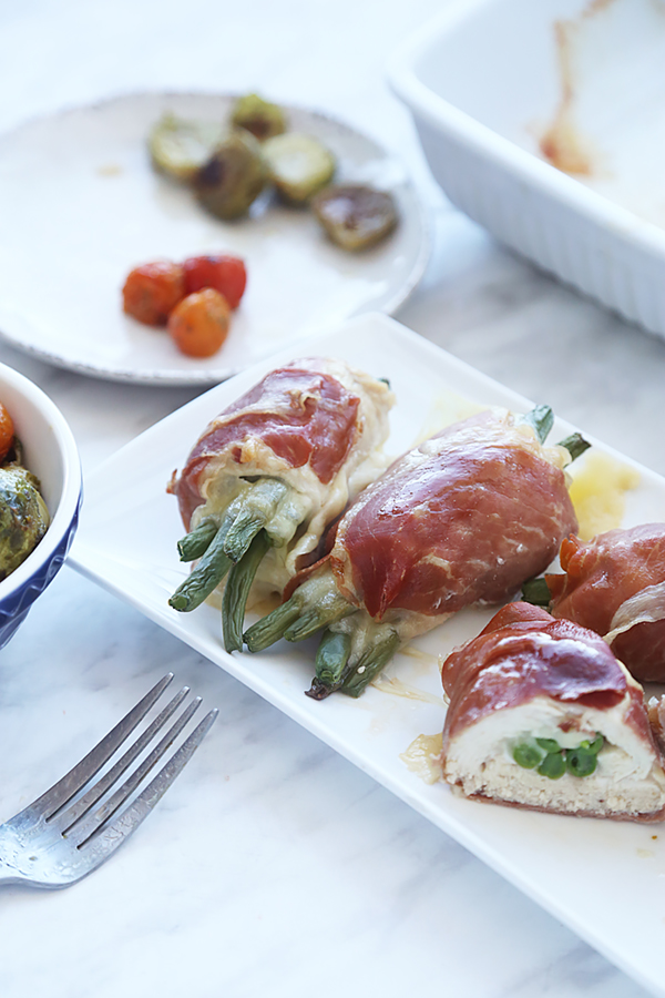 Golden prosciutto wrapped chicken breasts with melted swiss cheese and green beans on a white plate