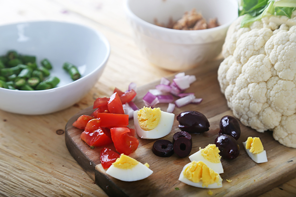 Diced tomatoes, hard-boiled egg, black olives, red onion, and green beans on a cutting board