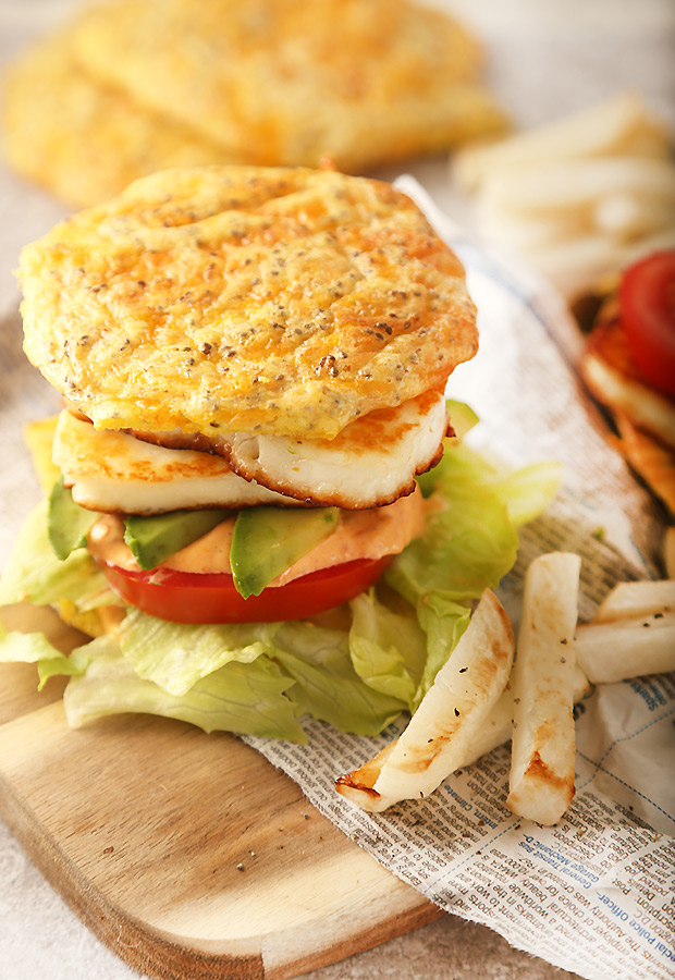 Assembled halloumi burger with lettuce, tomato, avocado, and red pepper sauce on a cheese bun with rutabaga fries