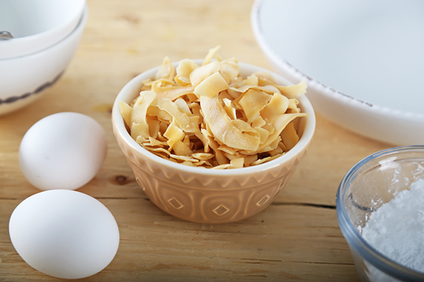 Toasted coconut flakes in a small bowl with eggs and powdered erythritol nearby