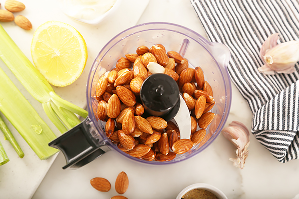 Whole soaked almonds in a food processor bowl with celery, lemon, and garlic alongside