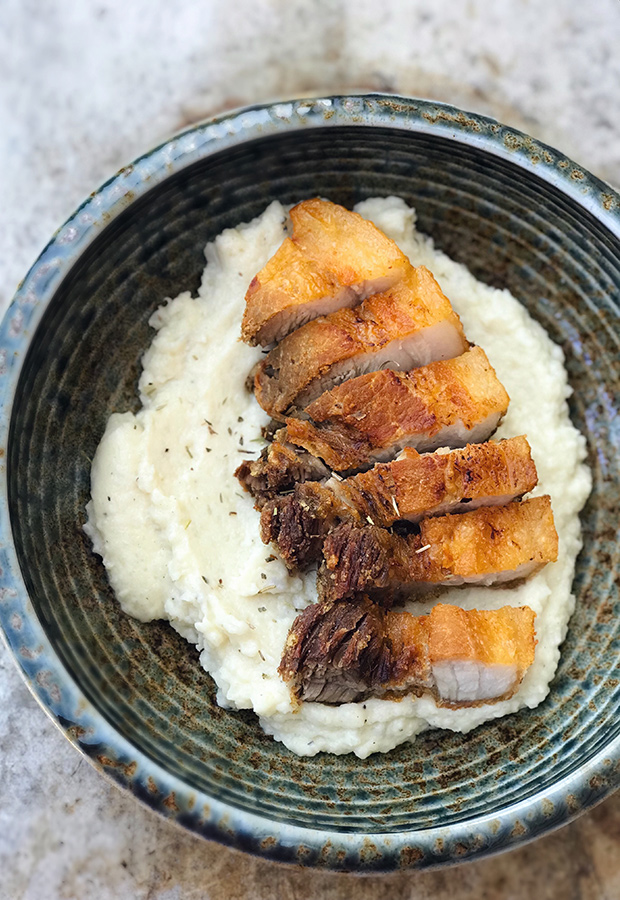 Sliced lechon kawali over herb cauliflower mash in a blue ceramic bowl