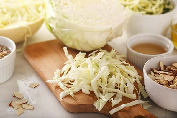 Shredded green cabbage on a wooden cutting board with sliced almonds and sunflower seeds nearby