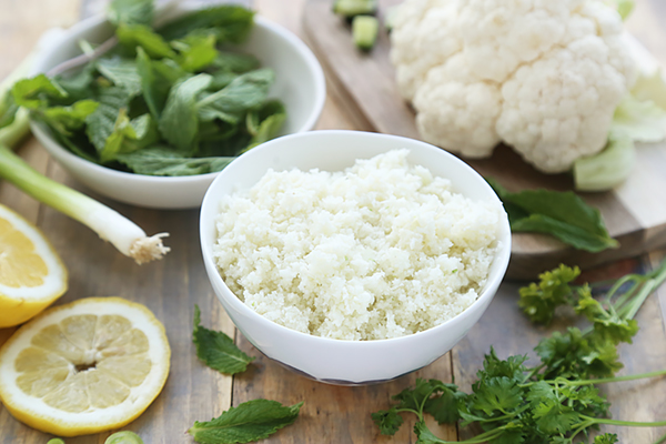 Riced cauliflower in a white bowl surrounded by fresh parsley, mint, lemon, and green onion